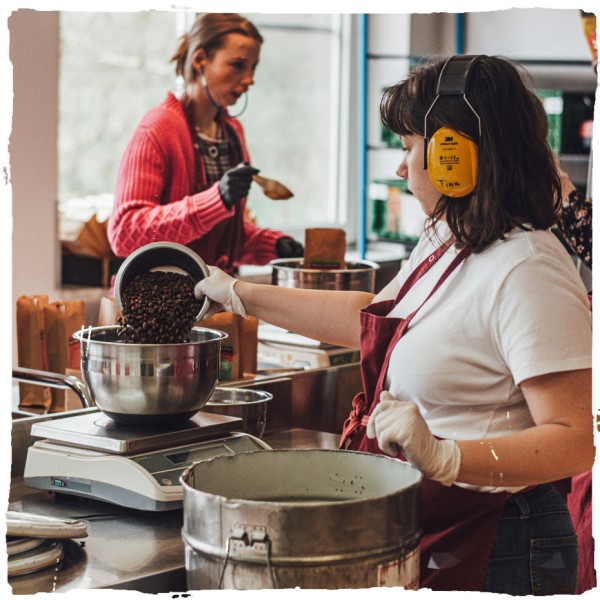 Eine Person im Vordergrund wiegt Kaffeebohnen ab. Im Hintergrund sieht man eine zweite Mitarbeiterin.