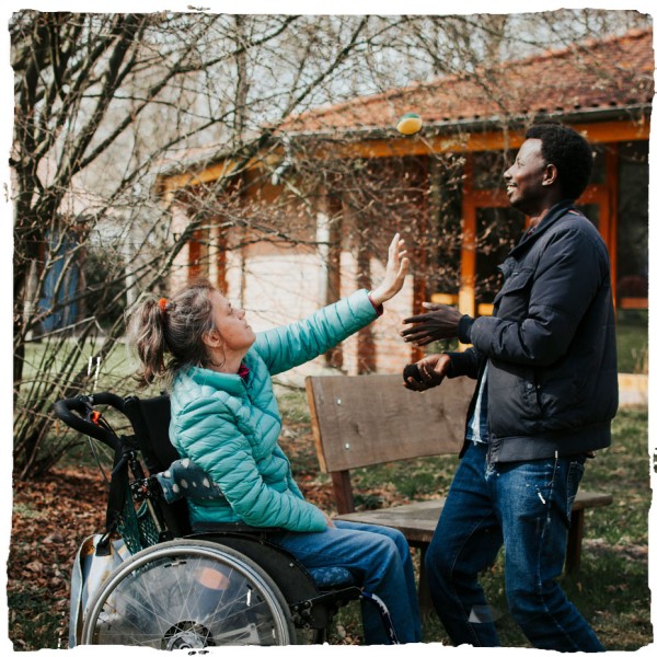 Ein stehender Mitarbeiter und eine Frau im Rollstuhl spielen gemeinsam mit einem Jonglierball.