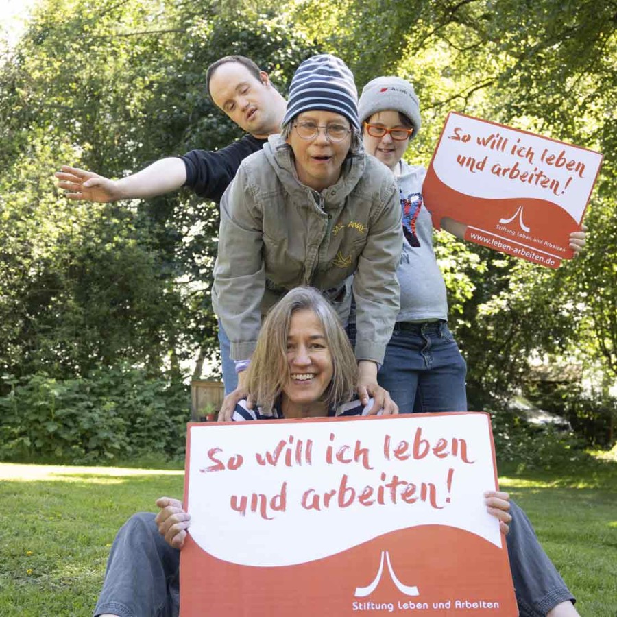 Vier Menschen der Stiftung Leben und Arbeiten stehen hintereinander und schauen uns aus dem Bild heraus an. Sie sind im Freien bei Sonnenschein. Sie halten Schilder auf denen Steht: So will ich leben und arbeiten.